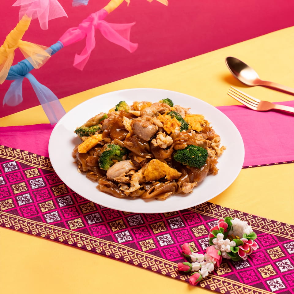 Pad See Ew with chicken, broccoli, and scrambled egg on a white plate with Thai decor.