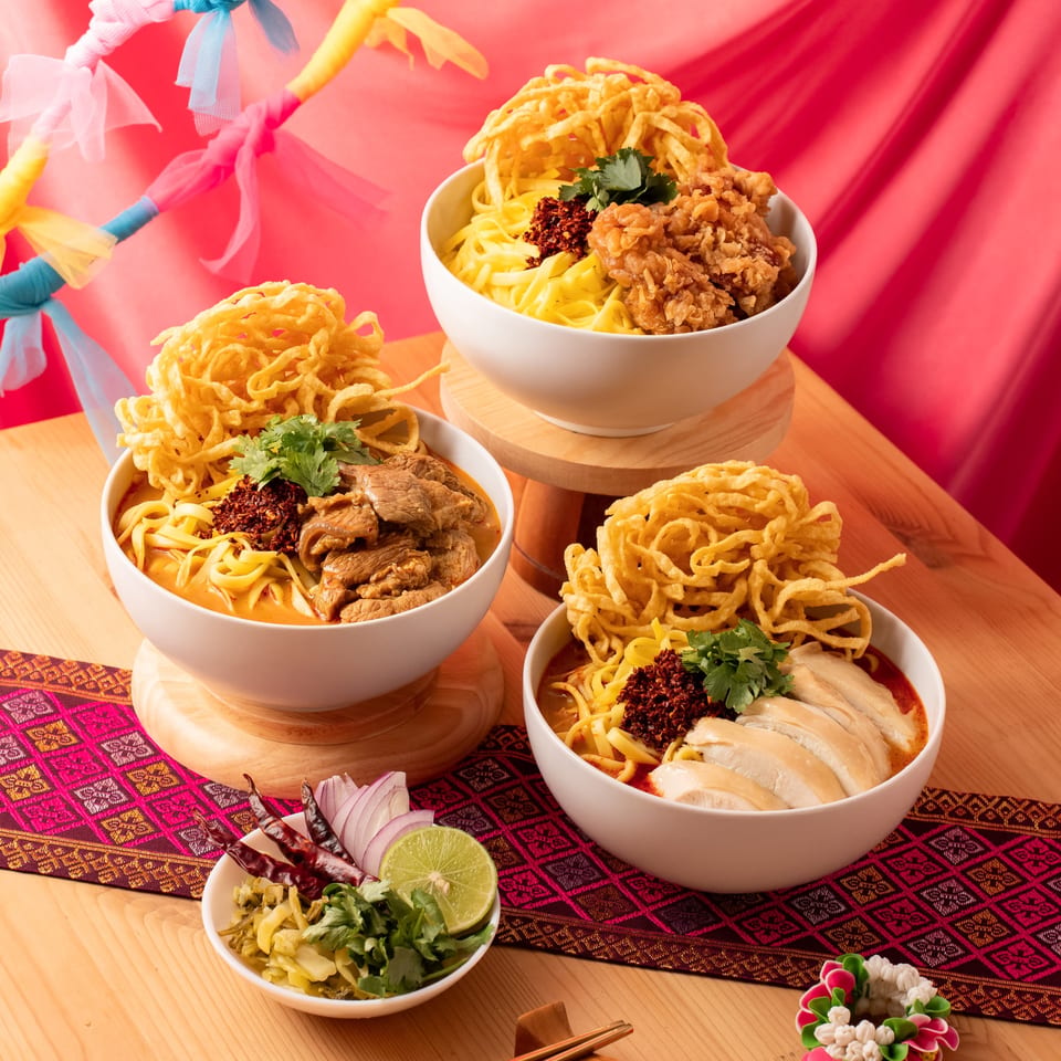 Three bowls of Khao Soi, with chicken, beef, and fried chicken, on a wooden table.