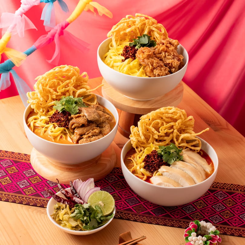 Three bowls of Khao Soi, with chicken, beef, and fried chicken, on a wooden table.