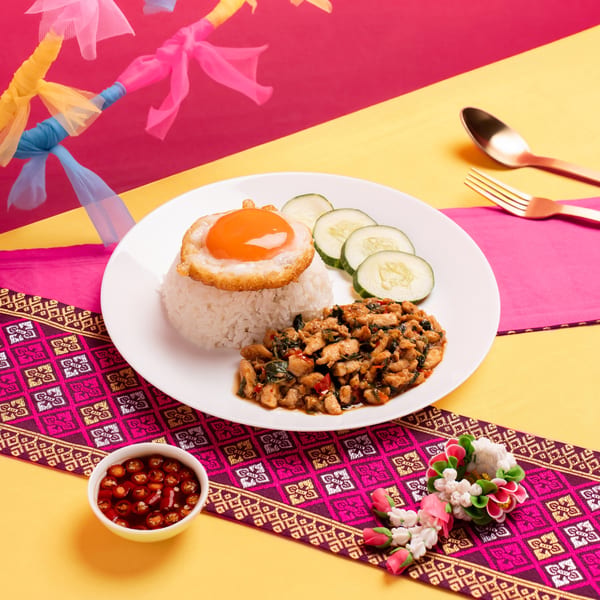 Thai basil chicken with fried egg, rice, and cucumber slices on a vibrant pink and yellow background.