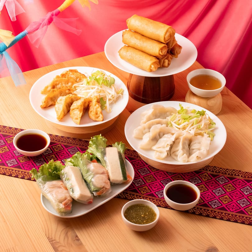 A wooden table laden with plates of spring rolls, dumplings, and fresh rolls, with dipping sauces.