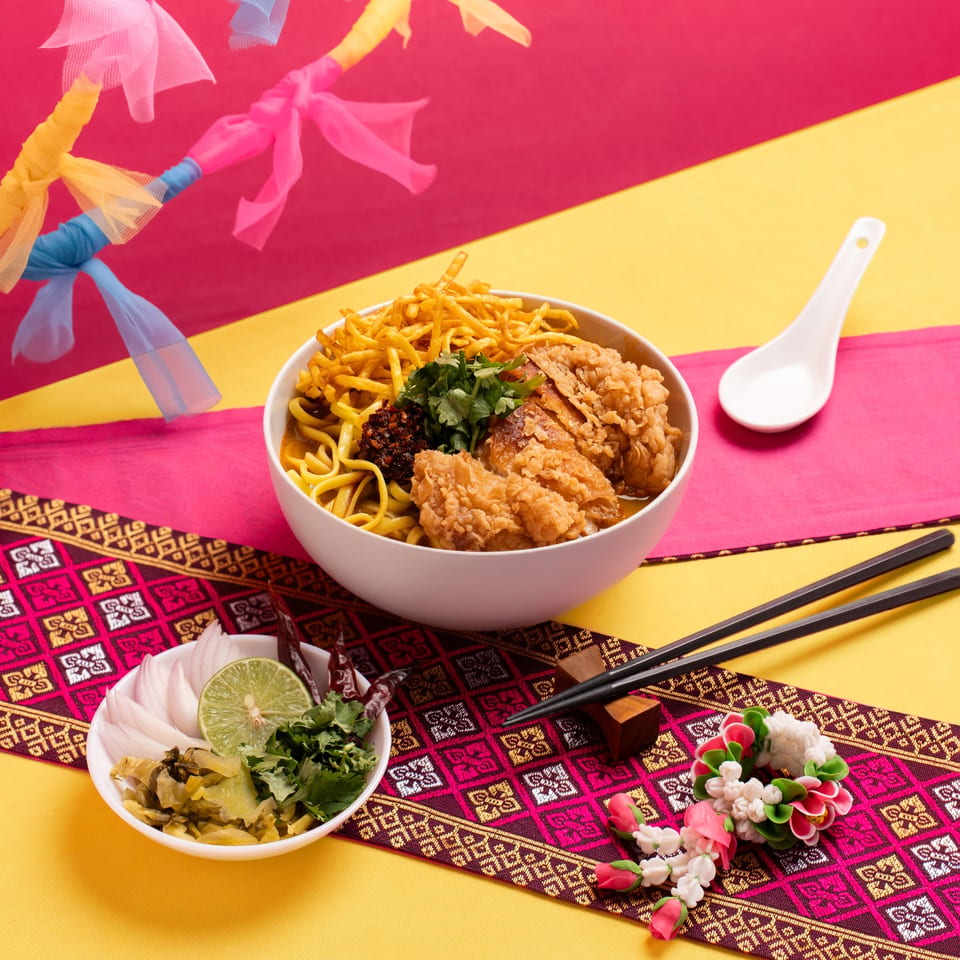 A bowl of Khao Soi with fried chicken, crispy noodles, and fresh herbs, served with condiments.