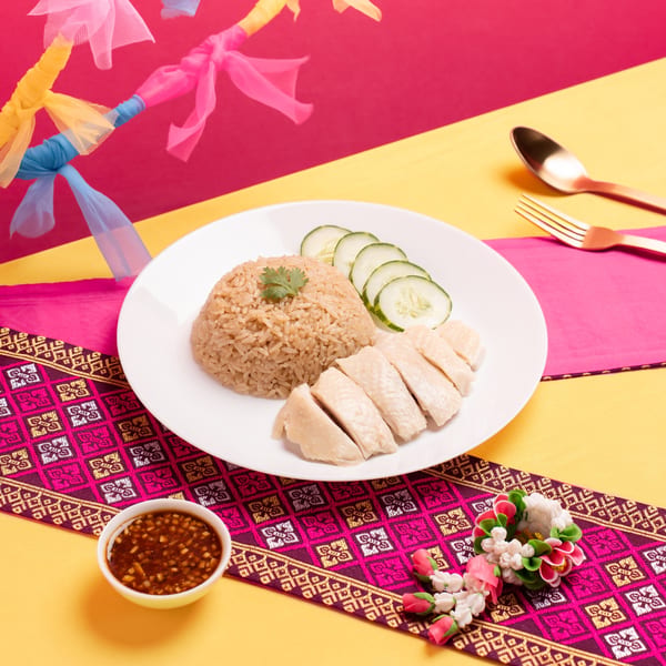 Hainanese chicken rice with sliced cucumbers, chili sauce, and decorative elements on a vibrant background.