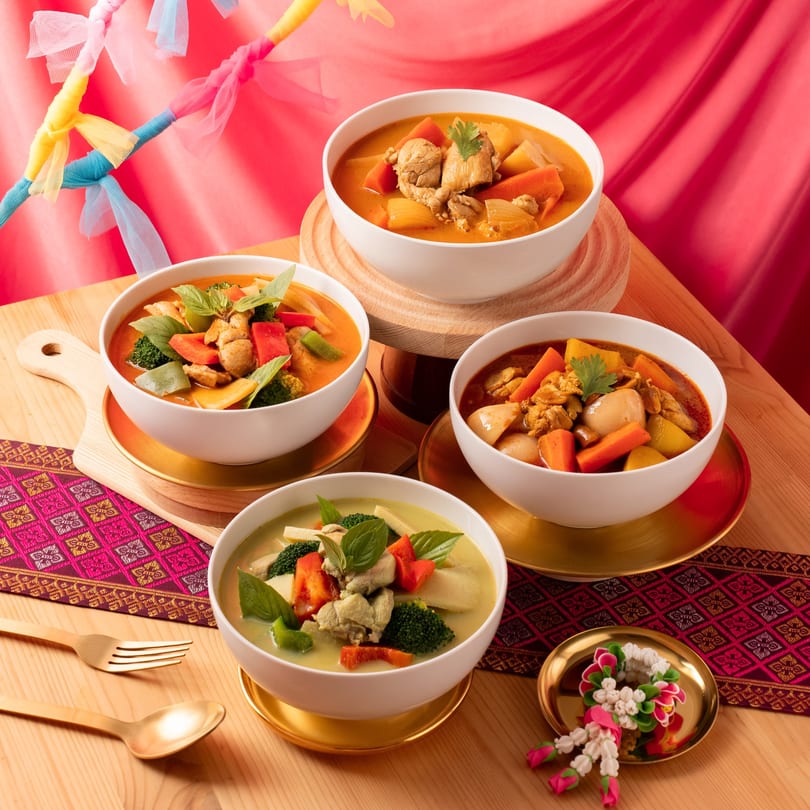 Four bowls of colorful Thai curries with chicken and vegetables on a wooden table with festive decor.
