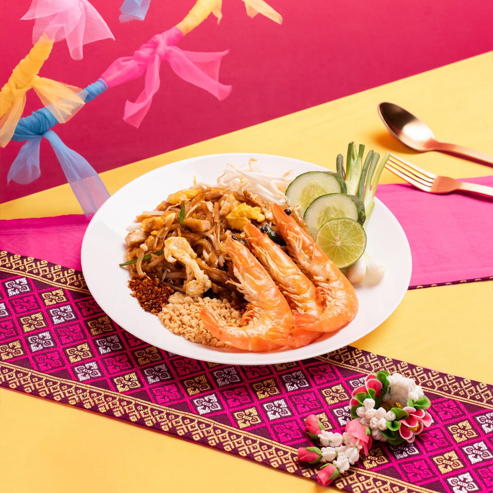 A plate of Pad Thai with shrimp, lime, and cucumber on a colorful table with decorative fabric.